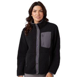A woman is wearing a black sherpa jacket with a high collar, snap-button closure, contrasting gray chest pocket, and side pockets.