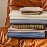 A stack of neatly folded bed sheets in various colors and patterns, including solid and striped designs, displayed on a brown leather chair.