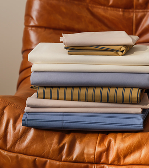 A stack of neatly folded bed sheets in various colors and patterns, including solid and striped designs, displayed on a brown leather chair.