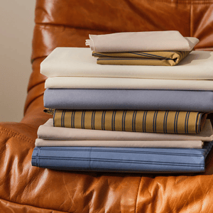A stack of neatly folded bed sheets in various colors and patterns, including solid and striped designs, displayed on a brown leather chair.