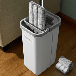 A grey self-wringing flat mop with a matching bucket for cleaning, featuring detachable microfiber pads, is placed next to rolled spare pads on a wooden floor.