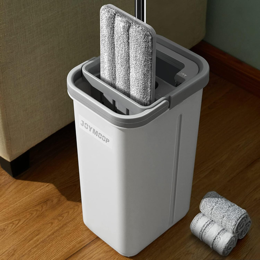 A grey self-wringing flat mop with a matching bucket for cleaning, featuring detachable microfiber pads, is placed next to rolled spare pads on a wooden floor.