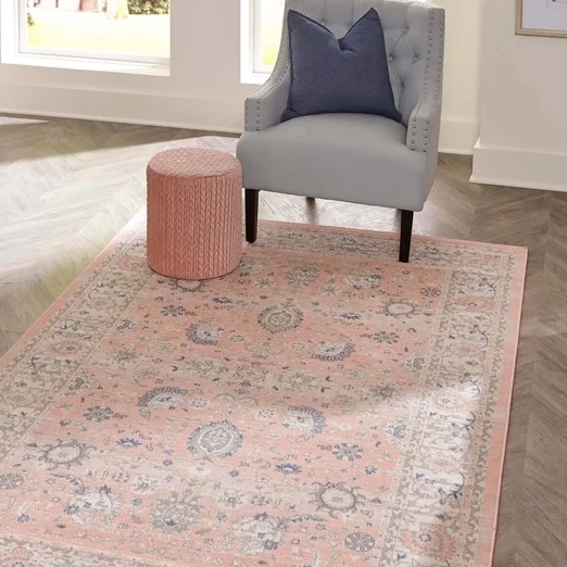 A gray wingback chair with a blue cushion, pink cylindrical ottoman, and a vintage-style pink area rug.