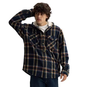 A plaid hooded shacket with sherpa lining, featuring chest pockets and button closures. The piece combines navy blue with traces of yellow, white, and red in the pattern.