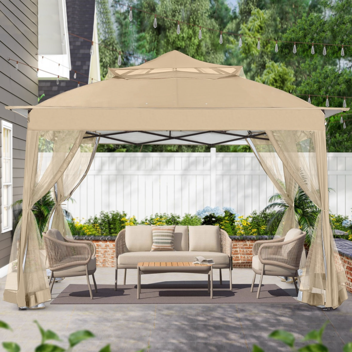 A beige outdoor gazebo with mesh curtains covers a patio set, including a sofa, two chairs, and a coffee table on a rug, situated on a stone patio with string lights and a garden backdrop.