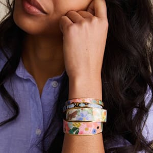 A person is wearing a set of colorful enamel cuffs with floral and butterfly designs, layered on their wrist. They are dressed in a light purple button-up shirt.