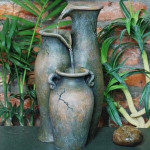 A decorative water fountain featuring three rustic, ceramic-style jugs with water cascading from the tallest to the smallest. Surrounded by lush greenery and set against a brick wall.