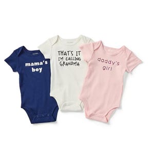 Three baby onesies with playful phrases: a blue one saying \“mama's boy,\“ a cream one with \“THAT'S IT I'M CALLING GRANDMA,\“ and a pink one with \“daddy's girl.\“