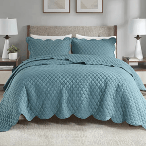 3pc Madison Park King Quilt Set includes a textured teal quilt with a scalloped edge and two matching pillow shams. The set decorates a bed with a clean and modern aesthetic, complemented by neutral bedside lamps and abstract wall art.