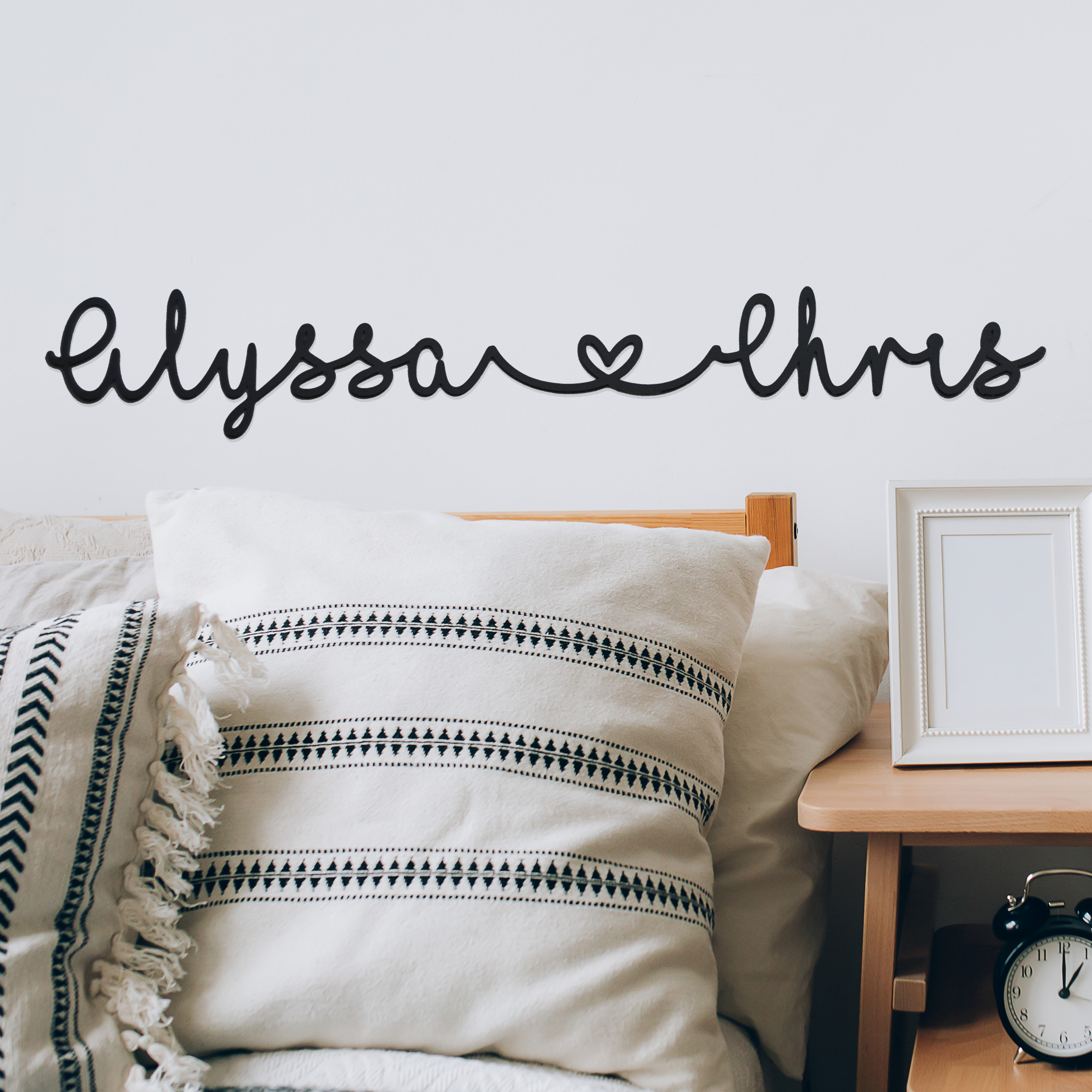 A decorative wall sign reads \“alyssa ♥ chris\“ above a bed with patterned cushions and a wooden side table featuring an empty photo frame and a small clock.
