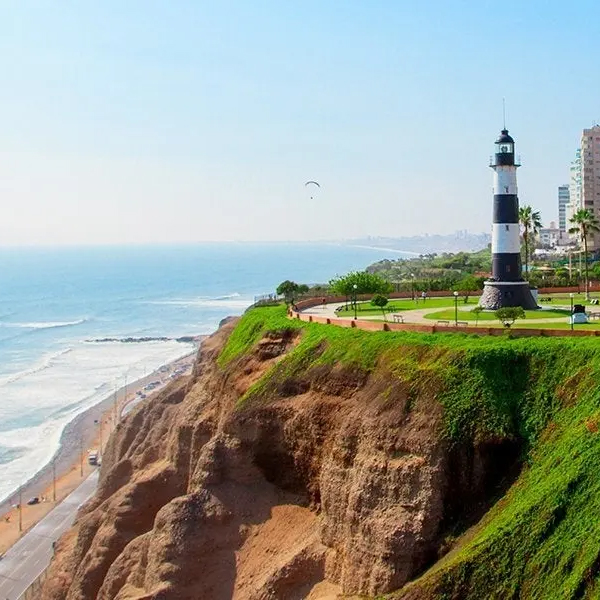 A scenic coastal view featuring a black-and-white lighthouse on a grassy cliff overlooking the ocean, with a paraglider in the sky and distant buildings in the background.