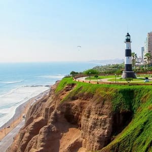 A scenic coastal view featuring a black-and-white lighthouse on a grassy cliff overlooking the ocean, with a paraglider in the sky and distant buildings in the background.