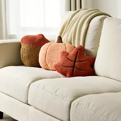 Three throw pillows on a couch: one resembles a brown acorn, another a pumpkin in shades of orange, and the third is a star-shaped pillow in a deeper rust color.