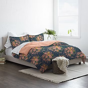 A twin-size comforter set with a dark floral pattern paired with a peach-colored sheet, accompanied by coordinating pillowcases, displayed on a bed with a white-fringed throw blanket by the side.
