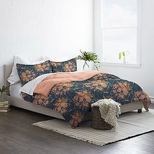 A twin-size comforter set with a dark floral pattern paired with a peach-colored sheet, accompanied by coordinating pillowcases, displayed on a bed with a white-fringed throw blanket by the side.