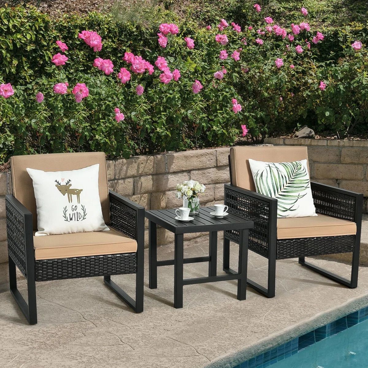 Two outdoor wicker chairs with beige cushions sit beside a small black table holding two cups and a vase with flowers, set against a backdrop of pink blooming roses.
