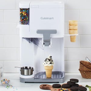 A Cuisinart soft serve ice cream maker with toppings dispenser, surrounded by cones, assorted toppings, and a bowl of cookies.