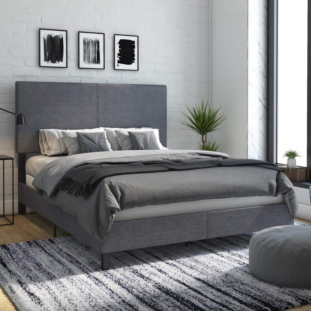 A modern bedroom features a grey-upholstered bed with a large headboard, layered with grey bedding and pillows. The room includes abstract art on the wall, a striped rug, a tall plant, and large windows allowing natural light.