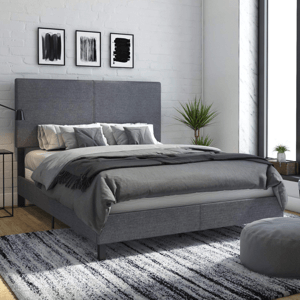 A modern bedroom features a grey-upholstered bed with a large headboard, layered with grey bedding and pillows. The room includes abstract art on the wall, a striped rug, a tall plant, and large windows allowing natural light.