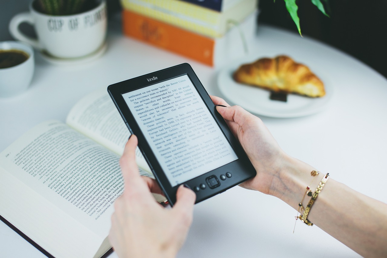 A person is holding an e-reader next to a printed book, coffee cup, and a croissant.