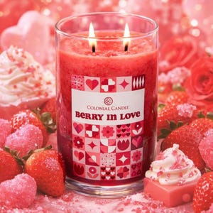 A lit Colonial Candle features the scent \“Berry in Love,\“ surrounded by strawberries, pink sugar decorations, and a pink frosted cupcake.