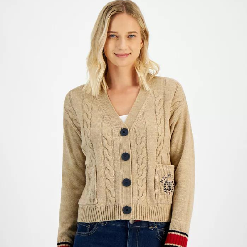 A beige cable knit cardigan by Tommy Hilfiger features ribbed cuffs with red and navy stripes, a v-neckline, front button fastening, and a patch pocket with a logo appliqué.