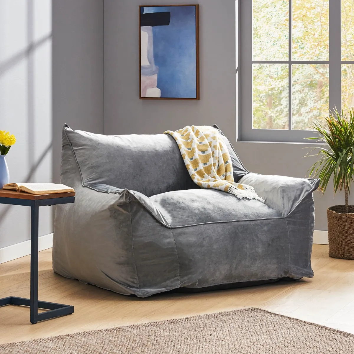 A gray bean bag chair with armrests is in a room with a small side table, a book, a patterned throw, a framed abstract painting, a potted plant, and a large window.