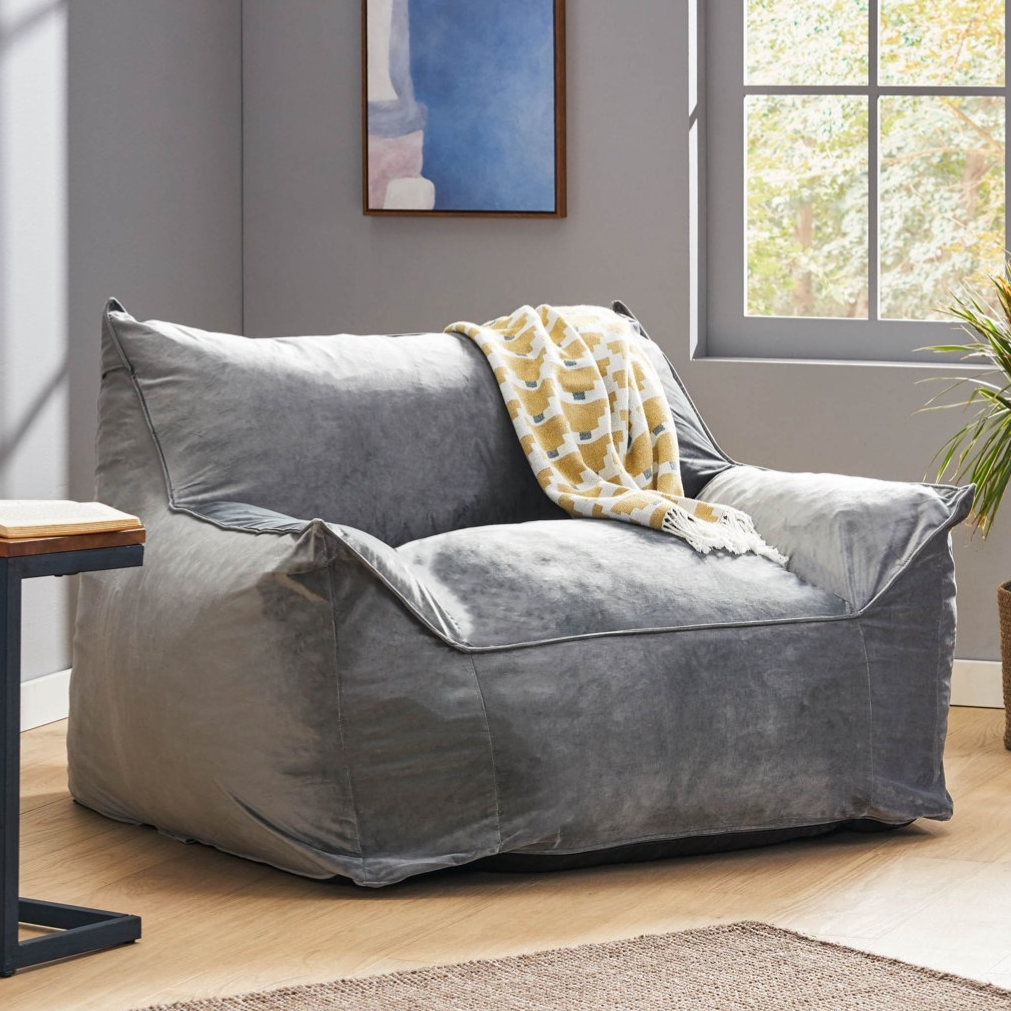 A gray bean bag chair with armrests is in a room with a small side table, a book, a patterned throw, a framed abstract painting, a potted plant, and a large window.
