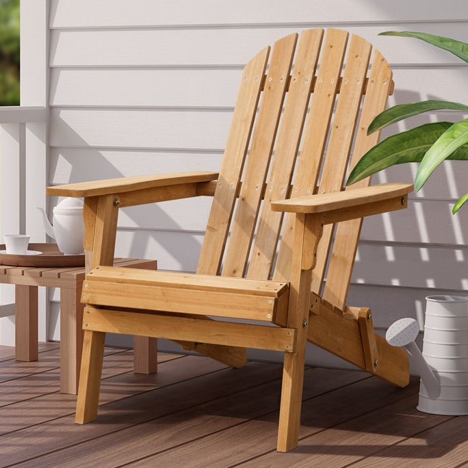A wooden folding Adirondack chair with classic slatted back and wide armrests is set on a deck with a small side table, teapot, cup, and a watering can nearby, next to a green plant.