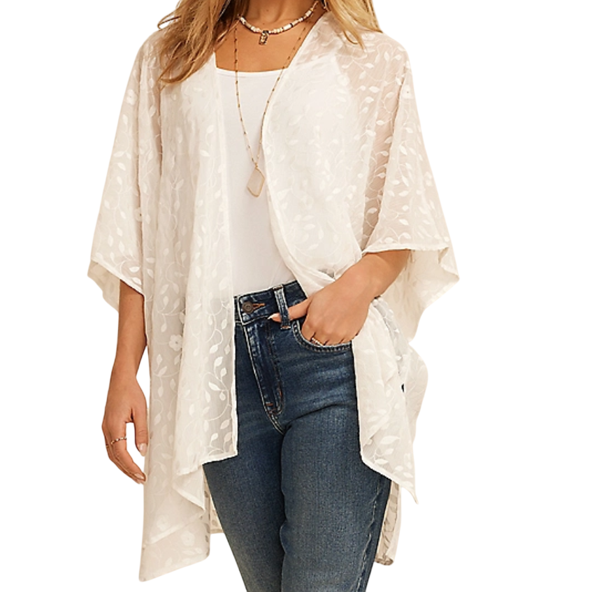A woman wears a white kimono with delicate floral patterns over a white top, paired with blue jeans. She's accessorized with layered necklaces.