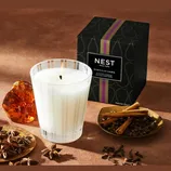 NEST New York Moroccan Amber scented candle with amber resin, star anise, cinnamon sticks, and cloves nearby. Packaging is black with a purple stripe and gold accents.