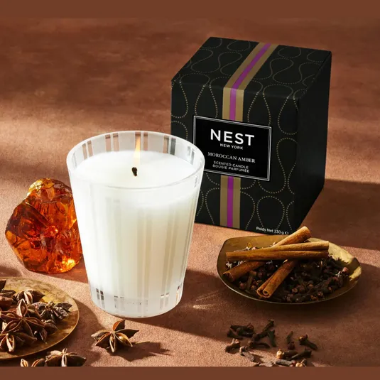 NEST New York Moroccan Amber scented candle with amber resin, star anise, cinnamon sticks, and cloves nearby. Packaging is black with a purple stripe and gold accents.