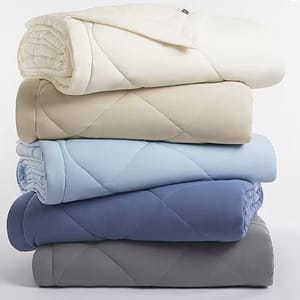 Stack of four quilted comforters in varying shades of cream, beige, light blue, and grey.