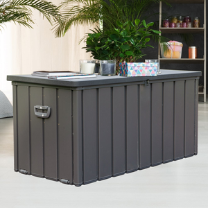 A dark gray 100-gallon deck box with vertical paneling, featuring a hydraulic lid for easy opening. It's used for outdoor storage and has a flat top used as a surface, with items like candles, a notebook, and a potted plant placed on it.