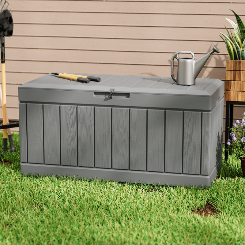 A gray outdoor storage bench with a shovel and trowel resting on top next to a metal watering can.