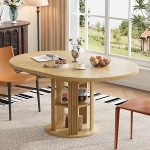 A wooden oval dining table with a central storage pedestal holds a magazine, a vase with flowers, and a fruit bowl. Two modern chairs, a floor lamp, and a sideboard with decorative items are in the background.