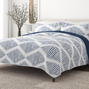 A bed with geometric-patterned blue and white bedding, accompanied by matching pillowcases. A vase with branches and a white lamp are on the bedside table, set against a textured rug.