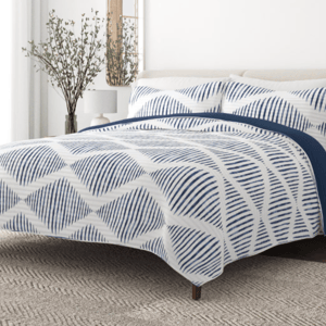 A bed with geometric-patterned blue and white bedding, accompanied by matching pillowcases. A vase with branches and a white lamp are on the bedside table, set against a textured rug.