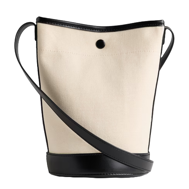 The Gap Factory Canvas Bucket Bag features a beige canvas body with a black leather base, trim, and strap. It includes a button closure at the top for security.