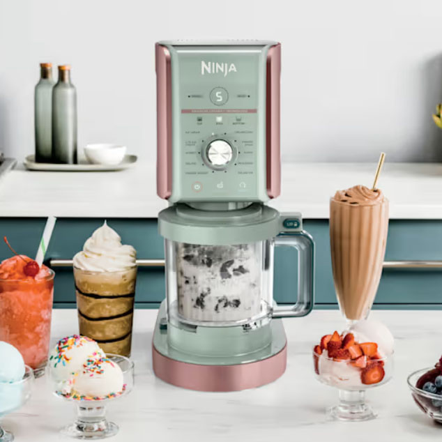 A Ninja ice cream maker is on a kitchen counter, surrounded by various desserts like milkshakes and ice cream sundaes adorned with toppings such as sprinkles, strawberries, and fruit.