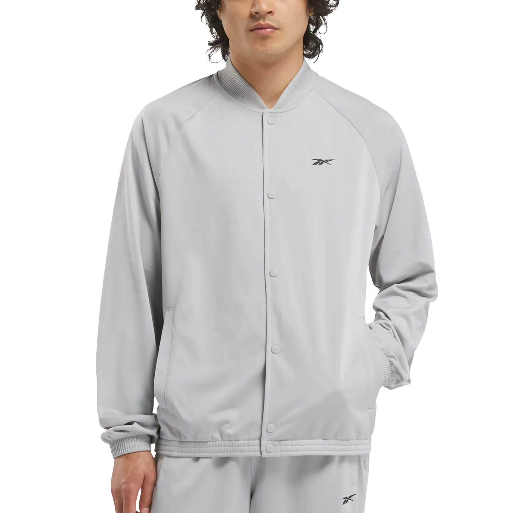 A man wears a light gray Reebok track jacket featuring a button-up front, ribbed cuffs, and a small logo on the chest, paired with matching track pants.