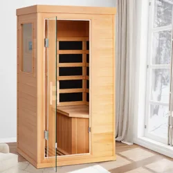 A wooden infrared sauna cabin with a glass door is placed in a room with snow visible outside the window.