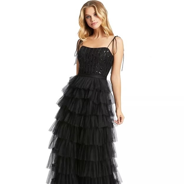 A woman wearing a full-length black dress with a tiered tulle skirt, thin shoulder straps, and a sequined bodice, creating an elegant and layered look.