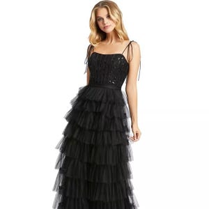 A woman wearing a full-length black dress with a tiered tulle skirt, thin shoulder straps, and a sequined bodice, creating an elegant and layered look.