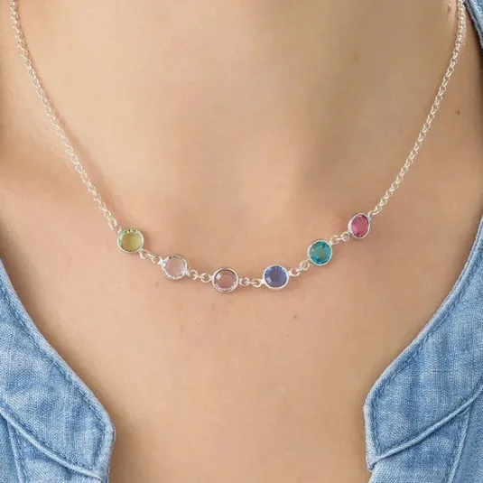 A delicate necklace with a series of colorful circular gemstones set in a chain, each stone in a different hue: green, pink, blue, turquoise, and red. The necklace is worn with a blue denim-style shirt.