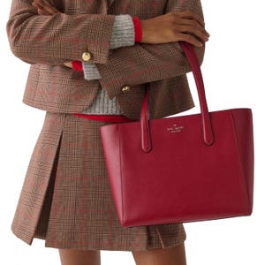 A person holds a red Kate Spade New York tote bag while wearing a plaid blazer and skirt, layered over a gray sweater.