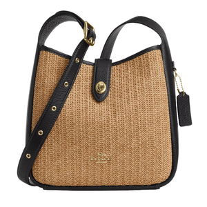A Coach shoulder bag with a woven beige fabric and black leather trim, featuring a gold-tone clasp and adjustable strap.