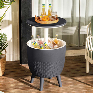 A round, elevated side table with a wicker-style finish features a built-in cooler compartment filled with ice and bottles. The tabletop above holds a tray with drinks and snacks.