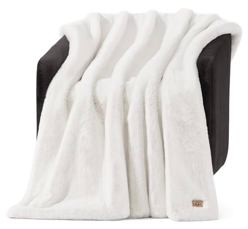A white faux fur throw blanket draped over a dark chair, featuring a small label with \“UGG\“ branding.