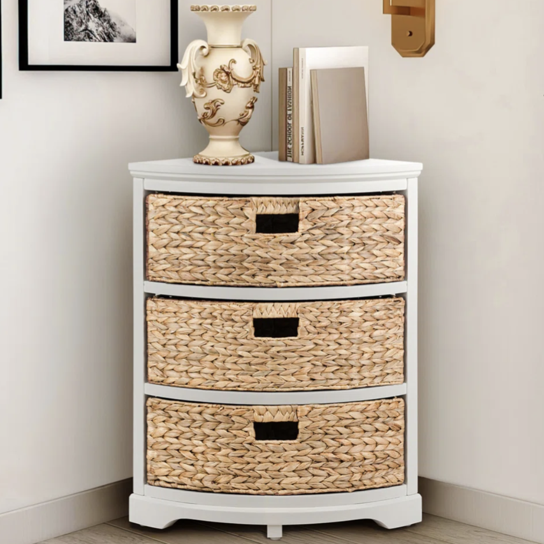 A corner chest with three woven drawers and a white top, decorated with a vase and books. It is placed beside framed art and a wall-mounted sconce.